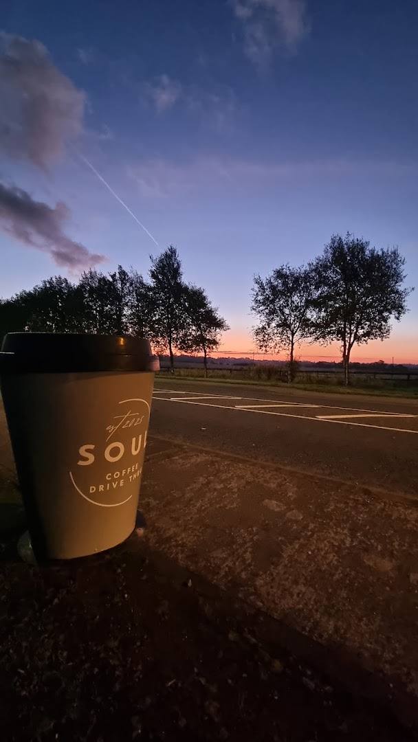 A branded coffee cup sits outdoors at dawn with a colorful sunrise over trees in the background.