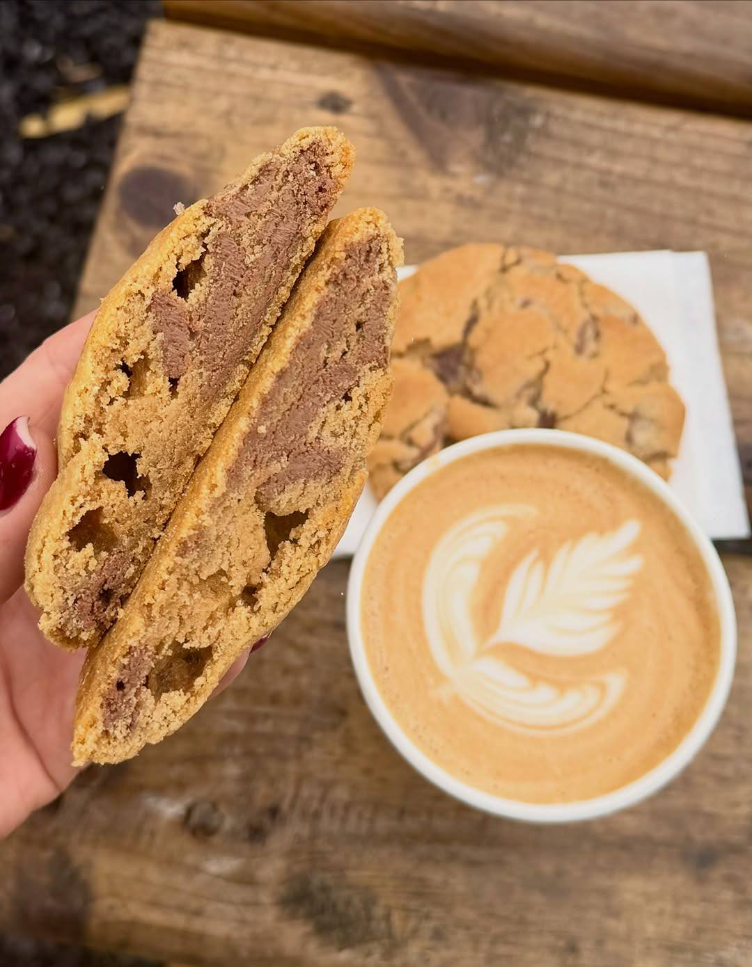 A hand holding a split chocolate cookie next to a latte with latte art on a wooden table